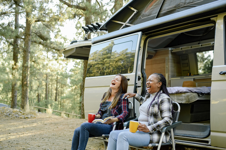 A younger and older middle aged woman laugh in front a van while camping. 